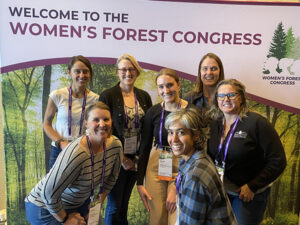 seven women smile at the camera in front of a large screen that says Welcome to the Women's Forest Congress.