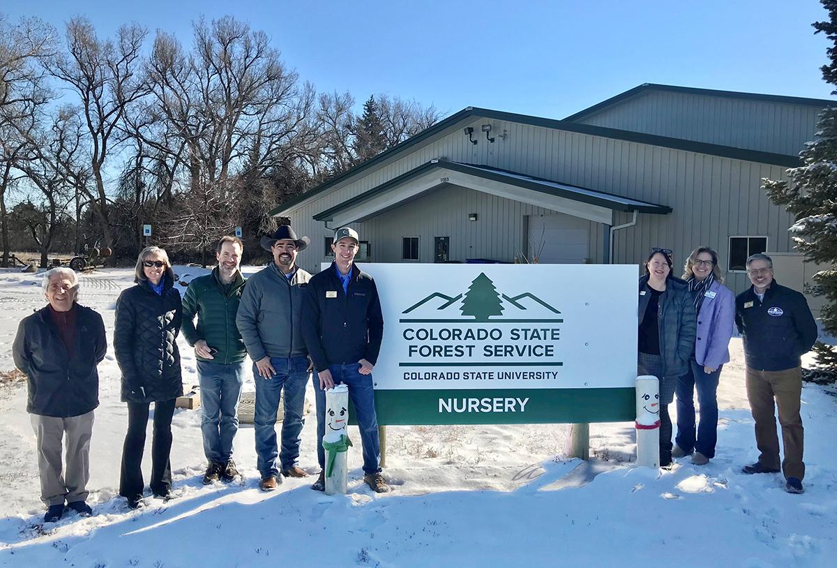 Group of 8 adults stand outside in a snowy yard next to a sign that reads Colorado State Forest Service Colorado State University Nursery.