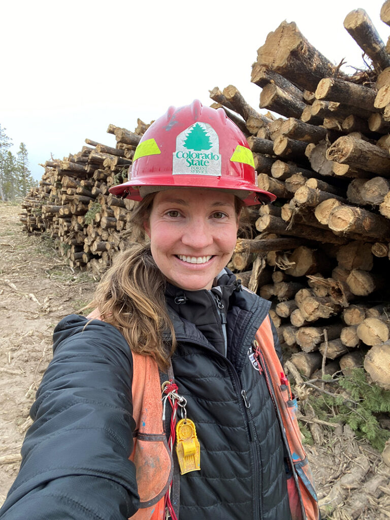 woman wearing hard hat and safety vest stands next to a large log pile.
