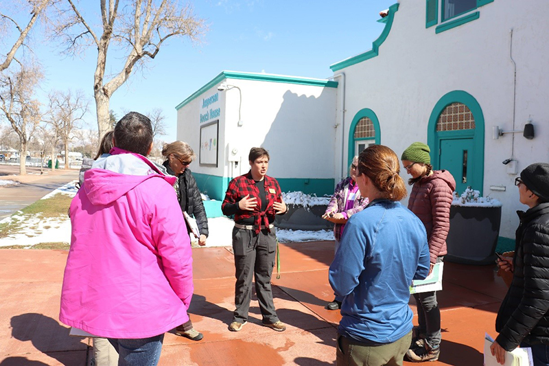 group of people stand in circle outside a white stucco building and listen to the person in the middle.