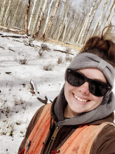 woman stands in snowy forest and smiles at the camera.