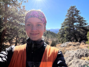 woman wears a safety vest while standing in a coniferous forest.