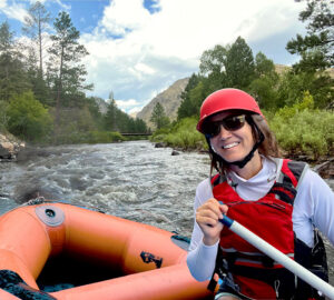 woman rafts down a river wearing a helmet and smiling at the camera. 