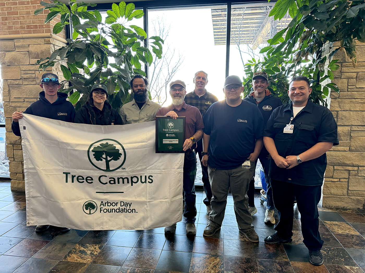 group of eight adults hold ups Tree Campus Arbor Day banner at San Luis Valley Health.